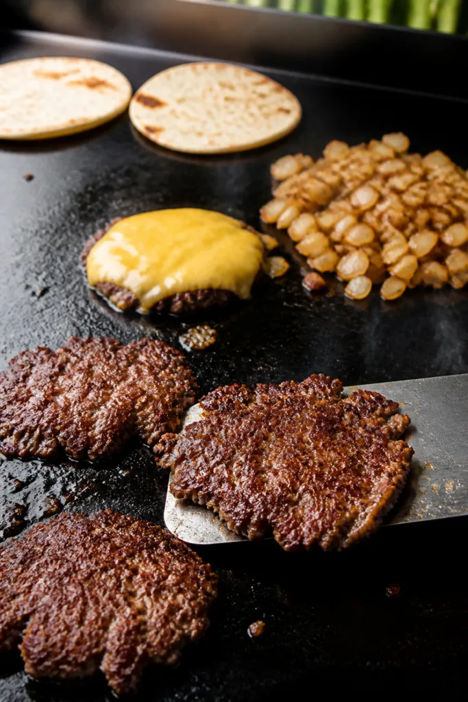 Cooking process close-up: On a Blackstone griddle, ultra-thin smash burger patties with deeply brown