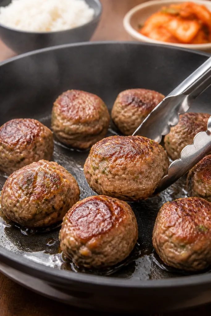 Cooking process close-up: Searing bulgogi meatballs in a large skillet over medium heat, golden-brow